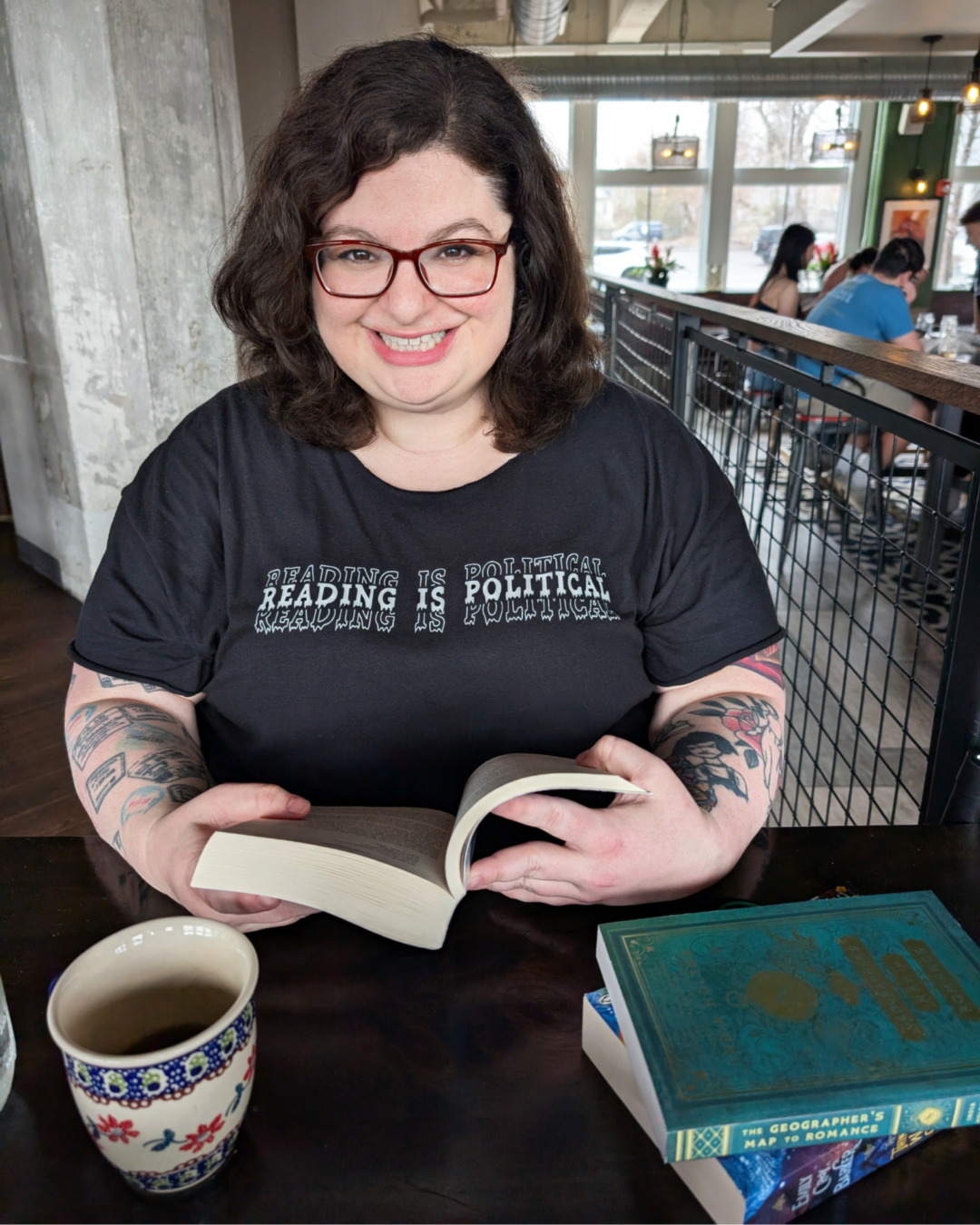 A woman with dark hair and glasses wearing a shirt that says "reading is political," while reading a book. 